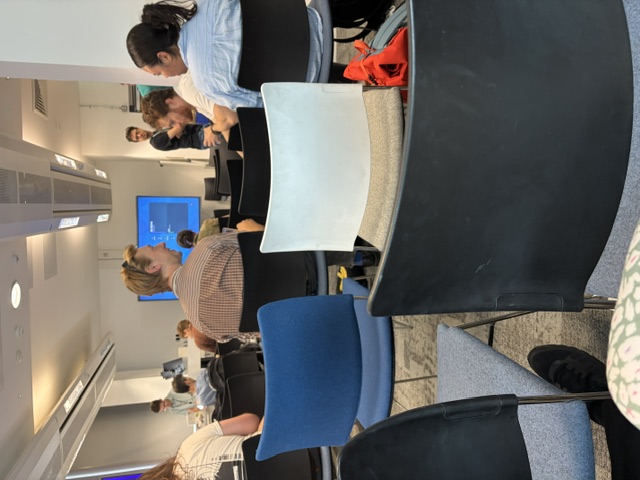 Image from the back of a room where people are sitting in chairs and chatting among themselves. Most of the chairs are empty and the front there's a person looking to the back of the room with their arms crossed.